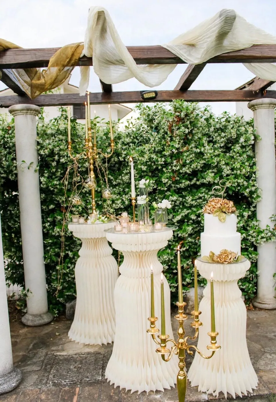 Pergolato con colonne e siepe di gelsomino, con torta nuziale bianca decorata da rose dorate tra i candelabri