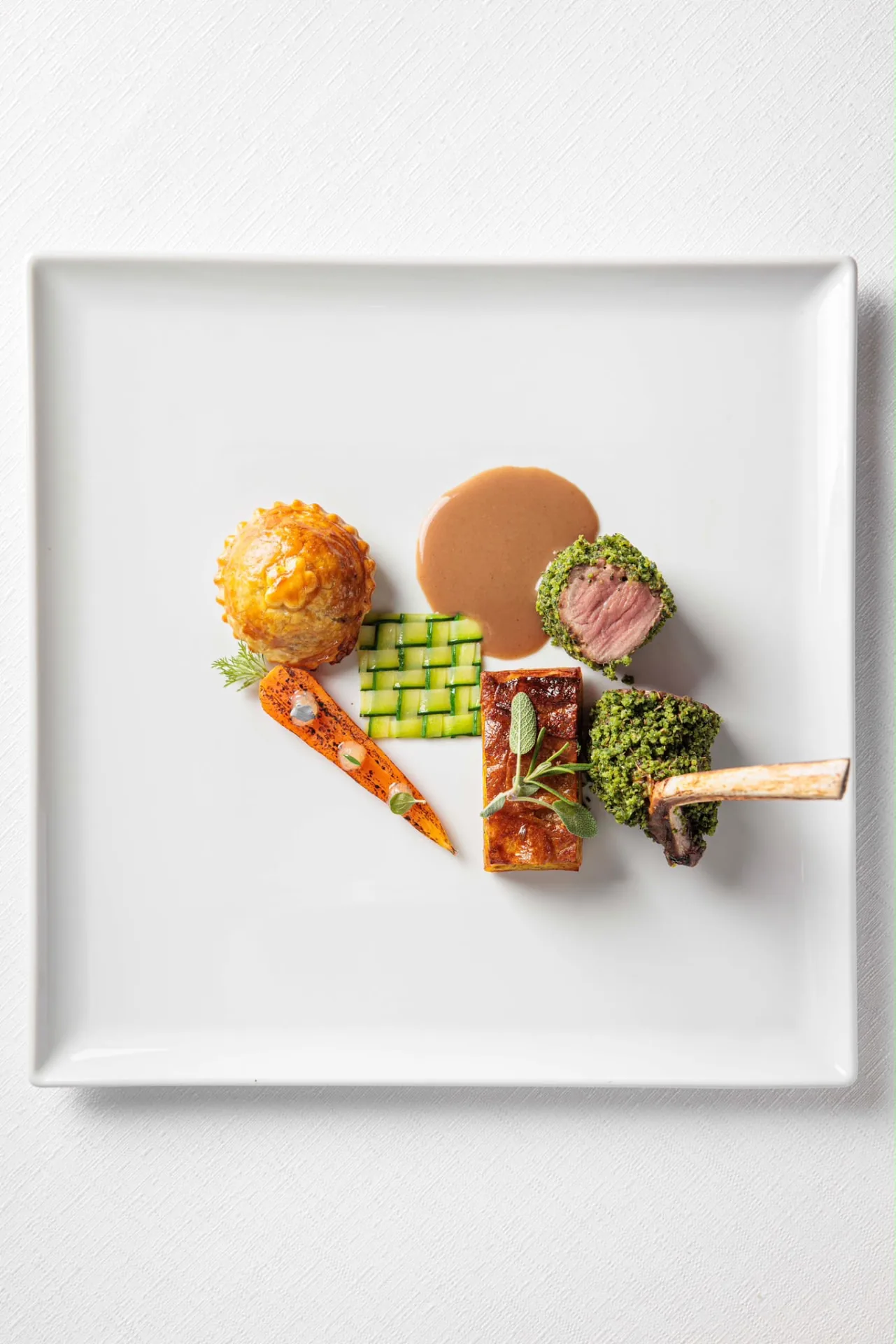 Agnello in crosta verde con costoletta, verdure e salsa su piatto quadrato bianco