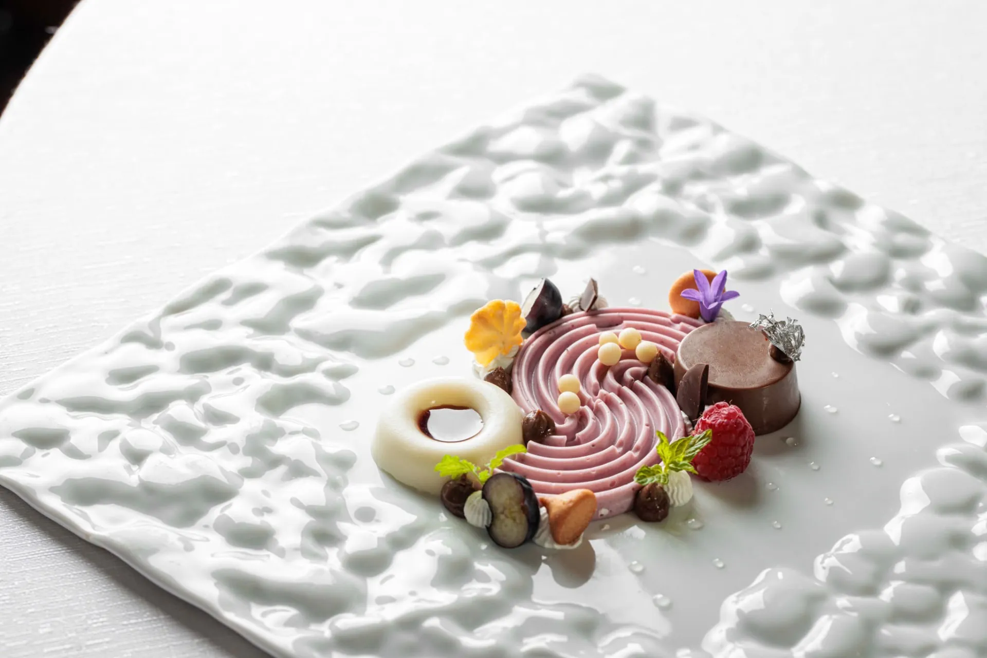 Dessert scomposto con spirale di mousse rosa, anello di panna cotta al caramello, cilindro al cioccolato con foglia d'argento, lamponi e mirtilli su piatto bianco materico